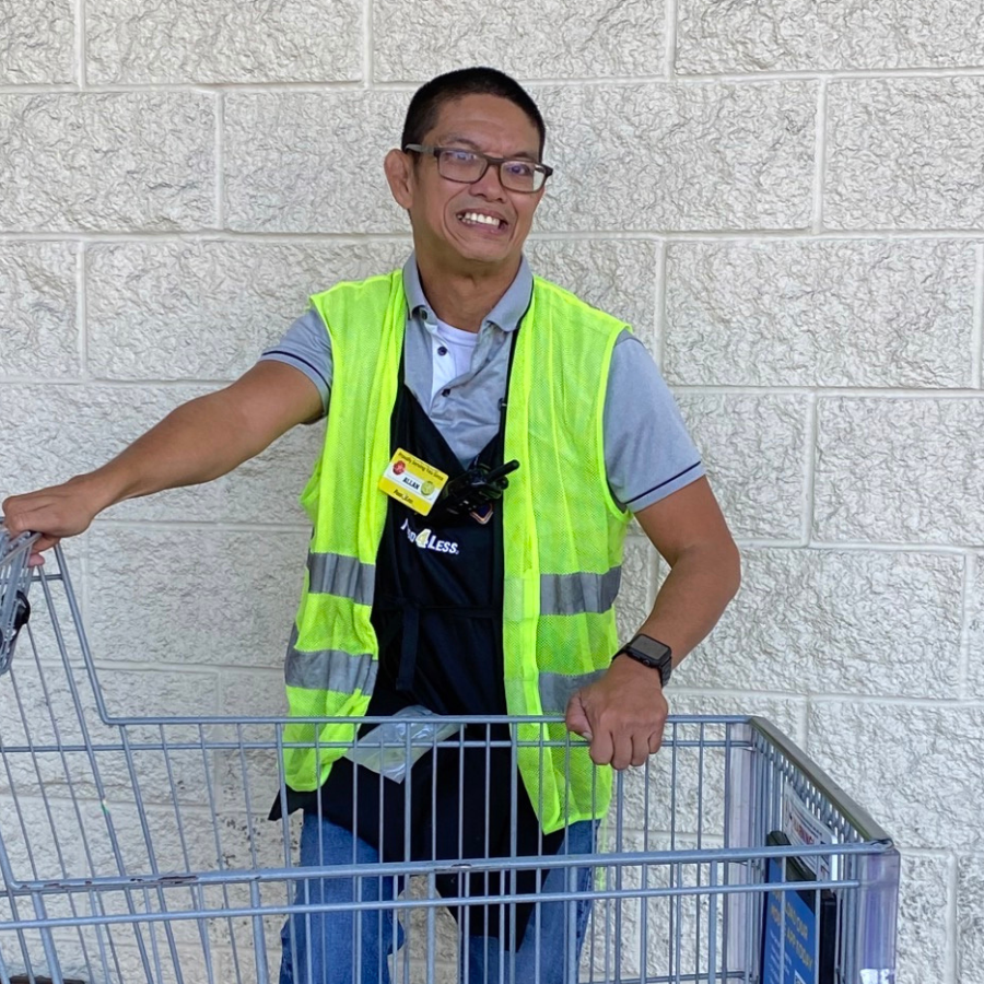 An Asian American man with short brown hair wearing glasses and a safety vest next to a grocery shopping cart